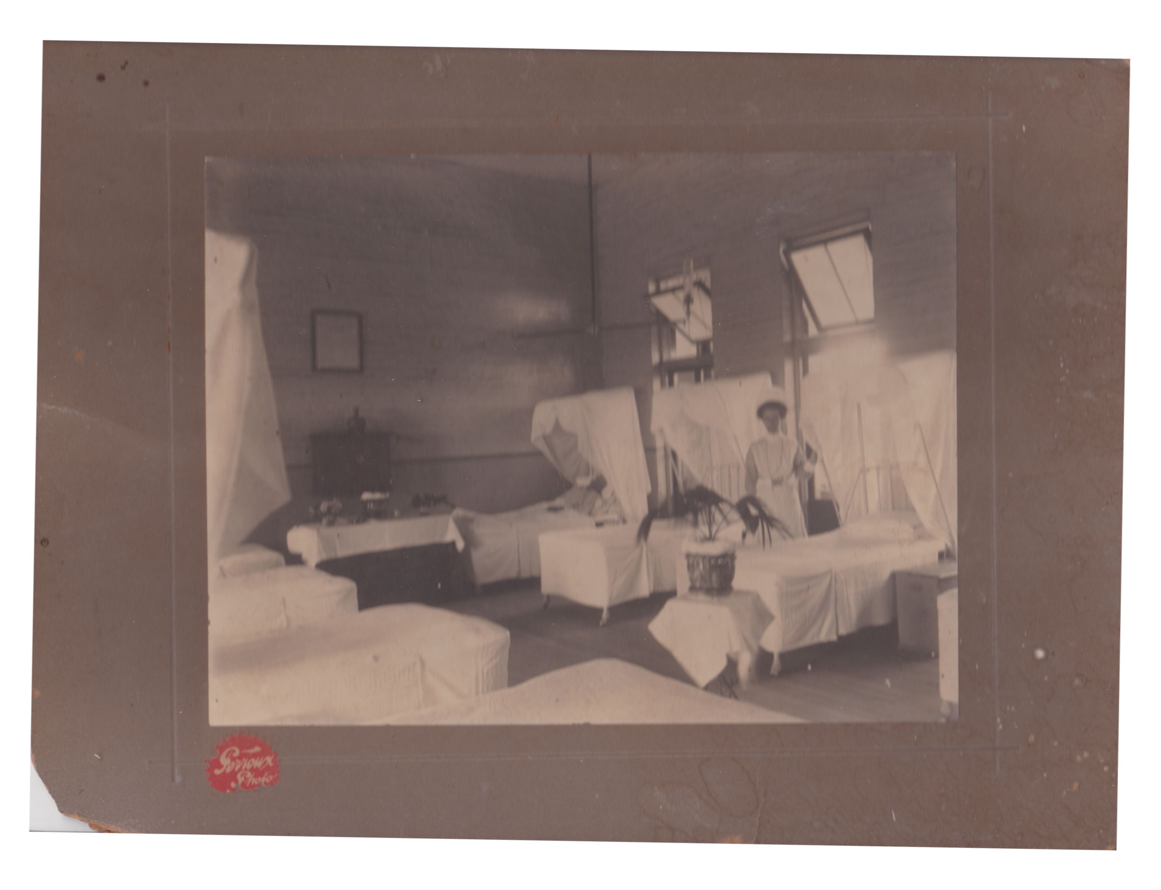 Black and white photograph of a ward inside Rockhampton General Hospital.  The room has high ceilings, and french doors, with open fanlight windows above them.  Seven beds are visible in the image, but there were probably more.  Each bed has a mosquito net.  There is a potted plant in the room, a picture on the wall, and what appears to be a sideboard against the wall with vases of flowers on it.  One patient lies in the far bed, and a nurse stands nearby.