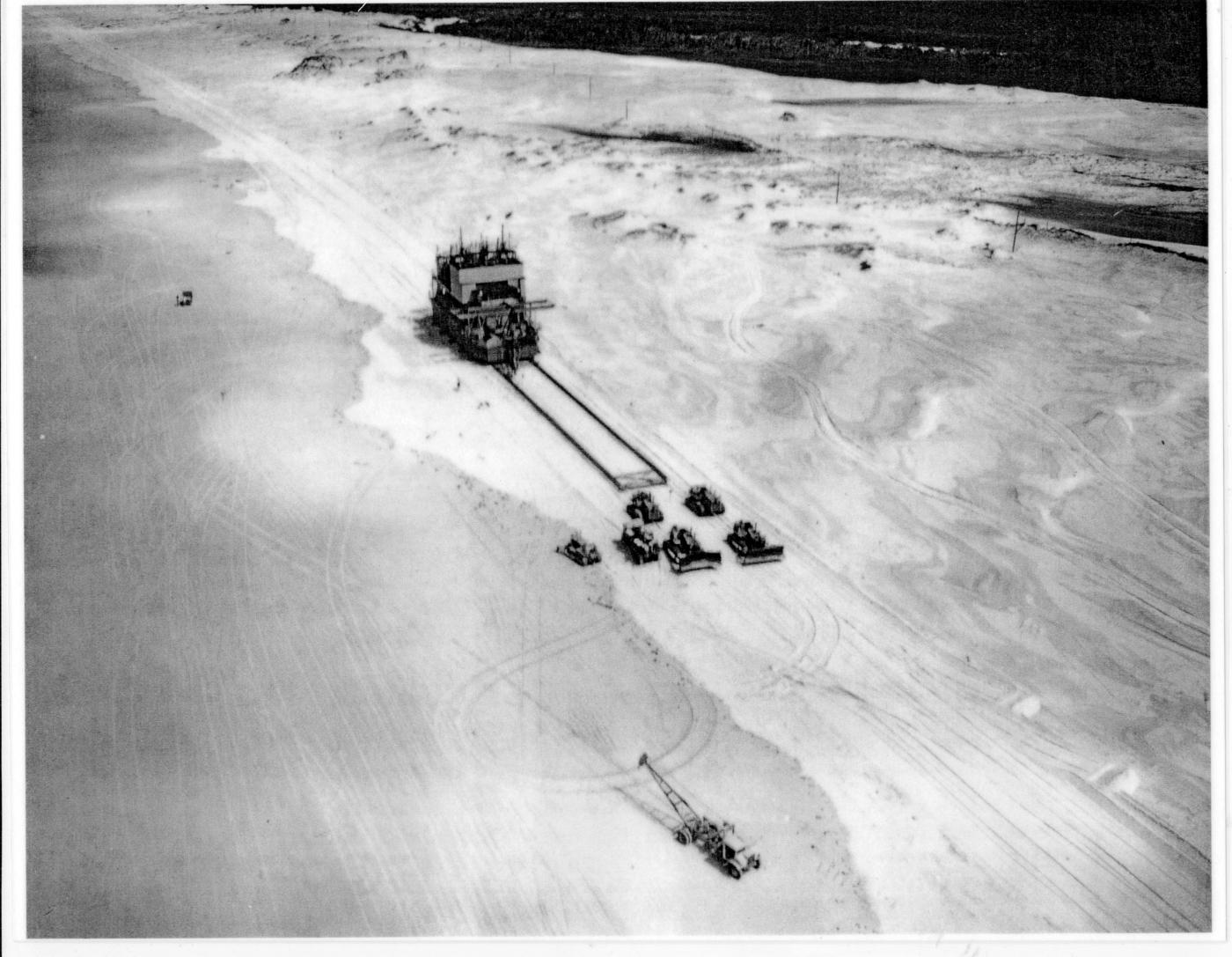 Moving a sandmining dredge along Main Beach on North Stradbroke Island, 1960s. Bulldozers are pulling the dredge, which is sliding on a constructed base.   Desc: Six bulldozers towing a mining dredge along a beach just above high tide mark. Beyond the sand dune is vegetation and an area of fresh water. The dunes are criss-crossed with tyre marks. 