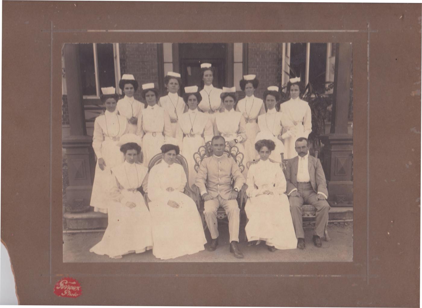 Black and white photograph taken at Rockhampton hospital showing newly graduated nurses, and staff members surrounding a doctor, who is seated centrally in the front row.  The nurses wear starched white uniforms and caps, and the doctor wears a light coloured suit. A second gentleman at the right of the front row may be the anaesthetist.