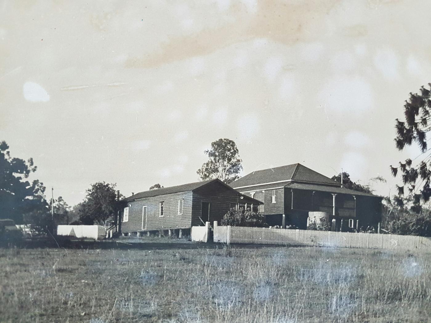 Black and white photograph of Kilburnie Homestead, taken approx 1950.   There is a single-storey detached kitchen and storeroom on the left, and the larger two-storey living areas of the homestead on the right.  A white picket fence and trees surround the structures.
