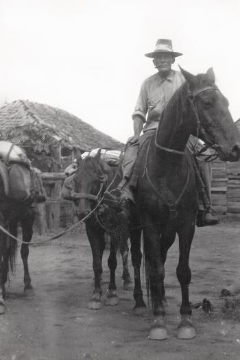 Black and White photograph of William Blair Campbell in the Kilburnie Saddling Yard with his bay horses.  He is riding one, and leading two packhorses loaded with gear.  He wears a wide-brimmed hat and work clothes, and is smoking a cigarette.