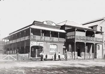 Black and white photograph of the Belle Vue Hotel, Rockhampton.  The Hotel is a two-storey brick building with a large veranda around the upper storey, and a veranda along the lower storey.  A large sign above the Hotel proclaims its name, and a large sign on the end of the lower storey veranda says "Alf Telford  Belle Vue Hotel".  A large painted sign on the window facing the street says "BAR".   Alf Telford, the proprietor, and his family and employees stand proudly on the street in front.