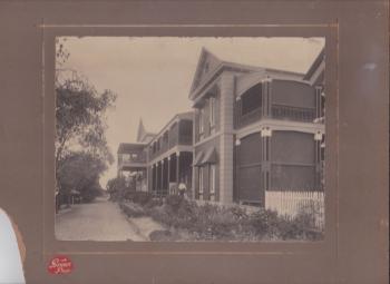 Black and white photograph of Rockhampton Hospital buildings, c.1910.  The two-storey buildings are brick and timber, with wide verandas upstairs and downstairs, surrounded by garden beds, with a gravel driveway., and white picket fencing