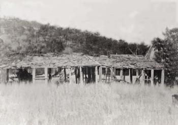 Black and white photograph of the ruins of the Rainbow Hotel.  The image shows a long, single-storey slab building in a state of decay, with large holes in the walls.  It is in the middle of a grassy paddock, with a hill in the background.